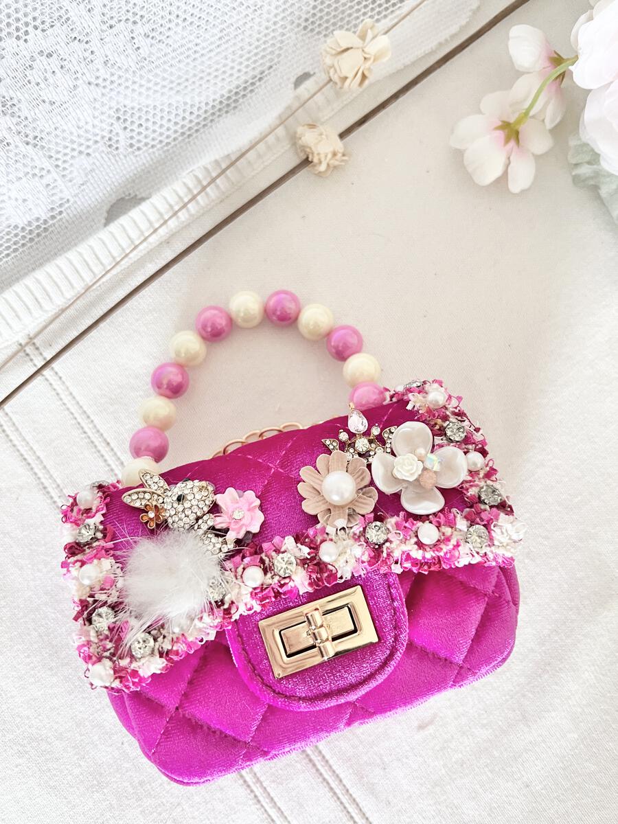 A bright pink quilted velvet handbag decorated with pink and white flowers, rhinestones, a white feather, and a beaded top handle, displayed on a light fabric background with decorative flowers.