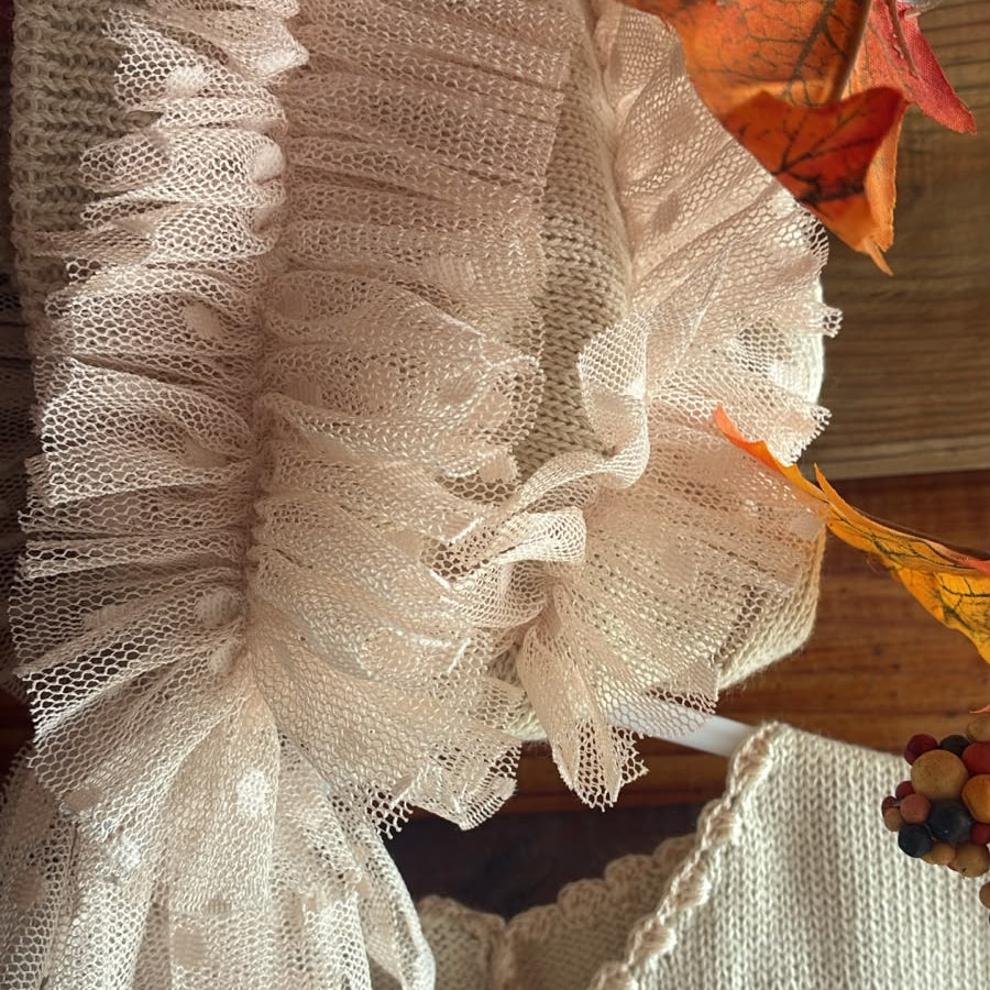 A close-up of a cream-colored knit garment with layered ruffle trim and a decorative beaded edge, styled with autumn leaves on a wooden surface.
