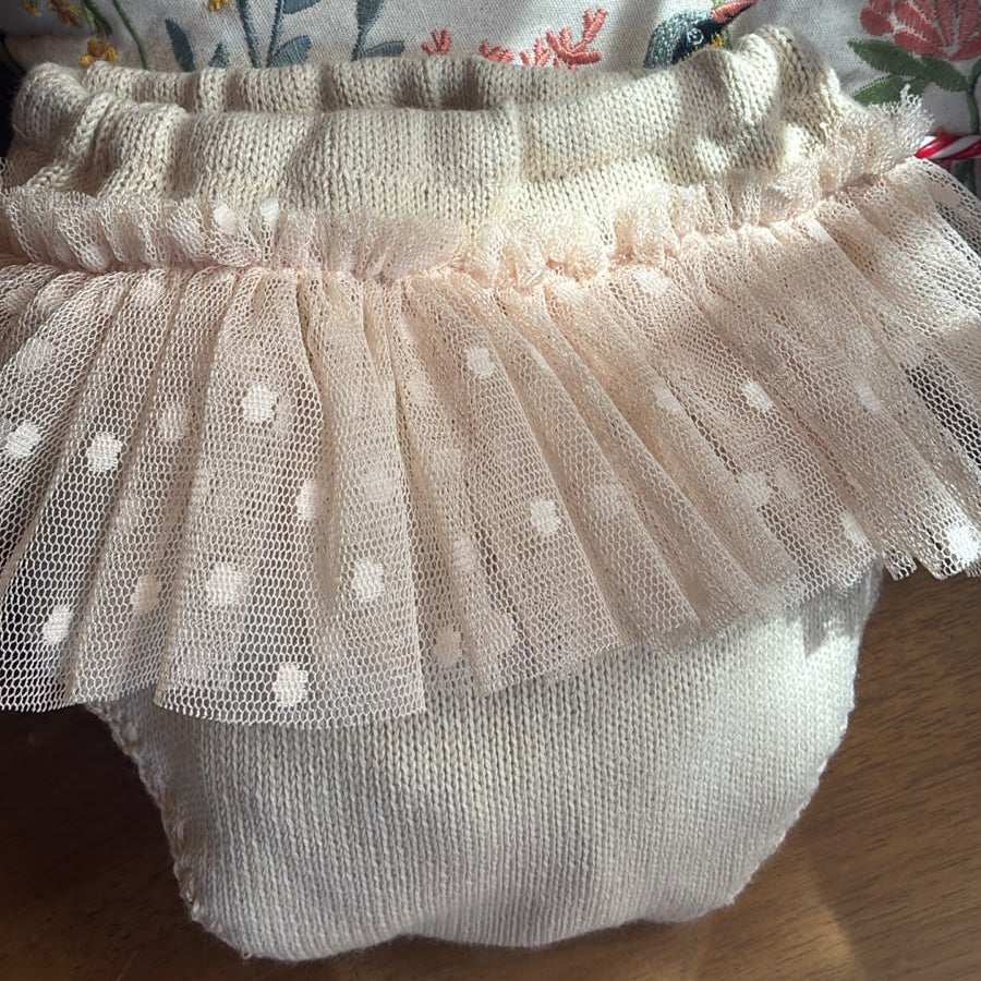 A close-up of a baby diaper cover or soft waist cover made of cream knit fabric with a layered ruffled tulle trim featuring white polka dots, resting on a floral embroidered fabric and wooden surface.