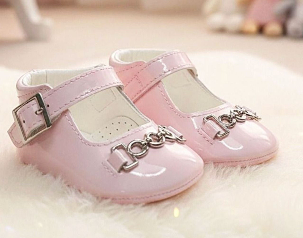 A pair of baby Mary Jane shoes in light pink patent leather with silver-tone hardware, displayed on a white faux fur surface.