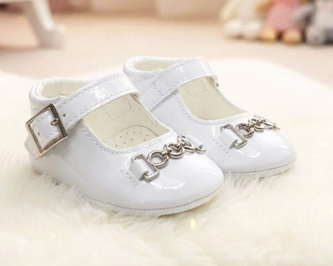 A pair of white patent leather baby Mary Jane shoes with silver buckle straps and decorative silver chain embellishments on the toes, resting on a white faux fur surface.