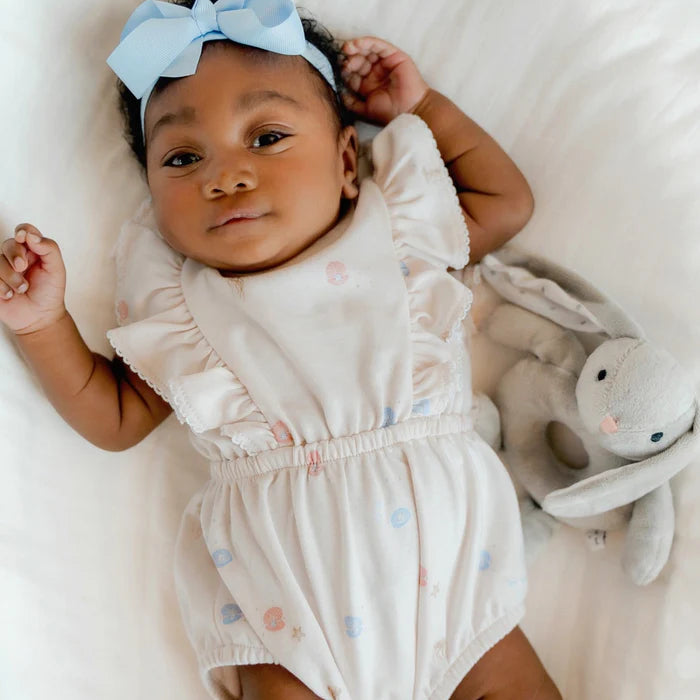 Baby in a white romper with colorful polka dots, wearing a blue bow headband, lying on a white blanket with a gray bunny toy.