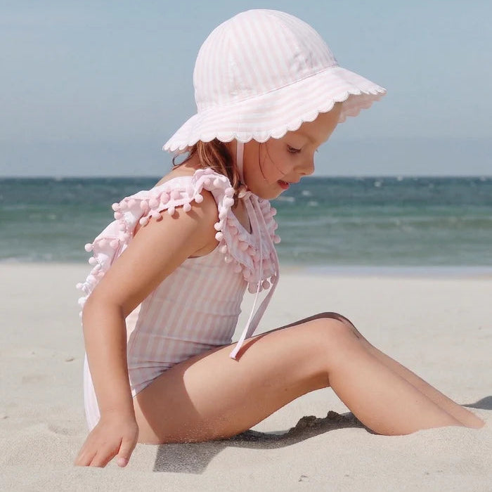 Child in a pink swimsuit and white sun hat sitting on a sandy beach with ocean in the background.